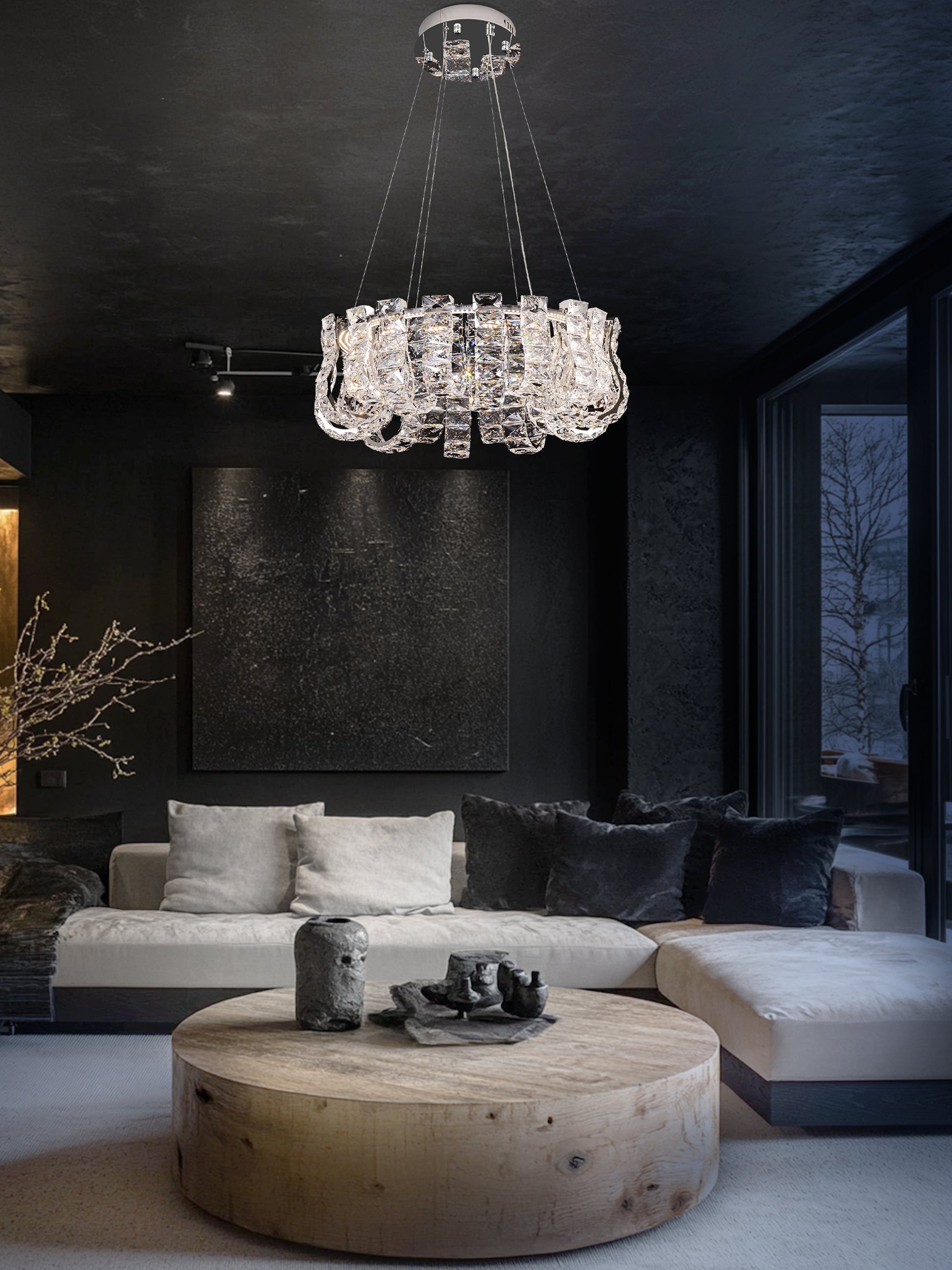 A modern crystal LED chandelier with a chrome finish hanging in a room with dark walls and a round wooden table in the foreground.