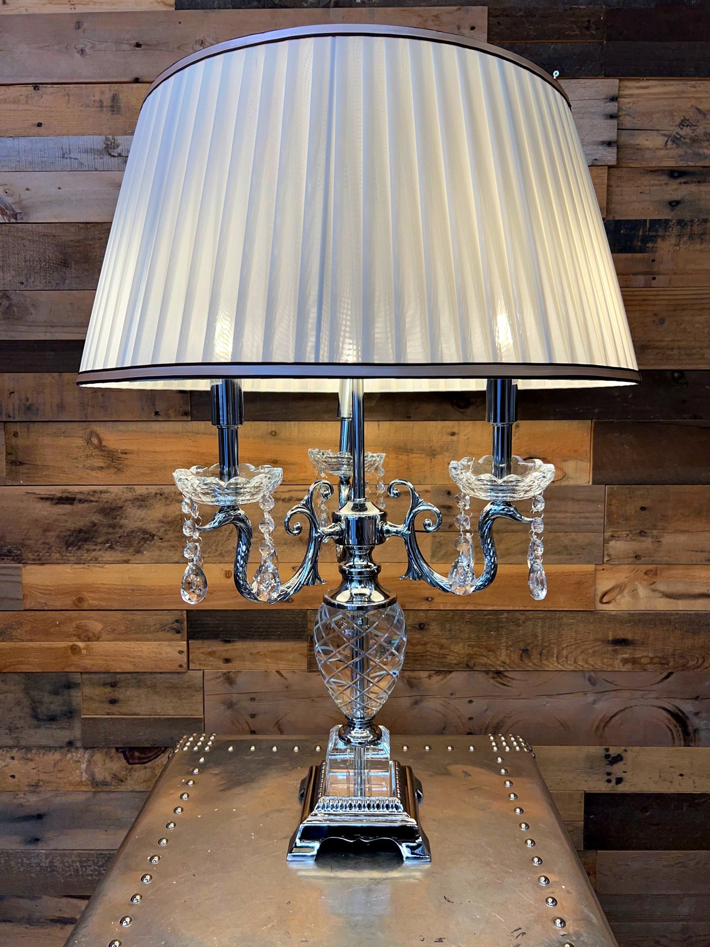 A traditional European crystal table lamp with a chrome finish and pleated shade. It has a sparkling faceted crystal base, chrome accents, and a brown-beige trim on the shade.